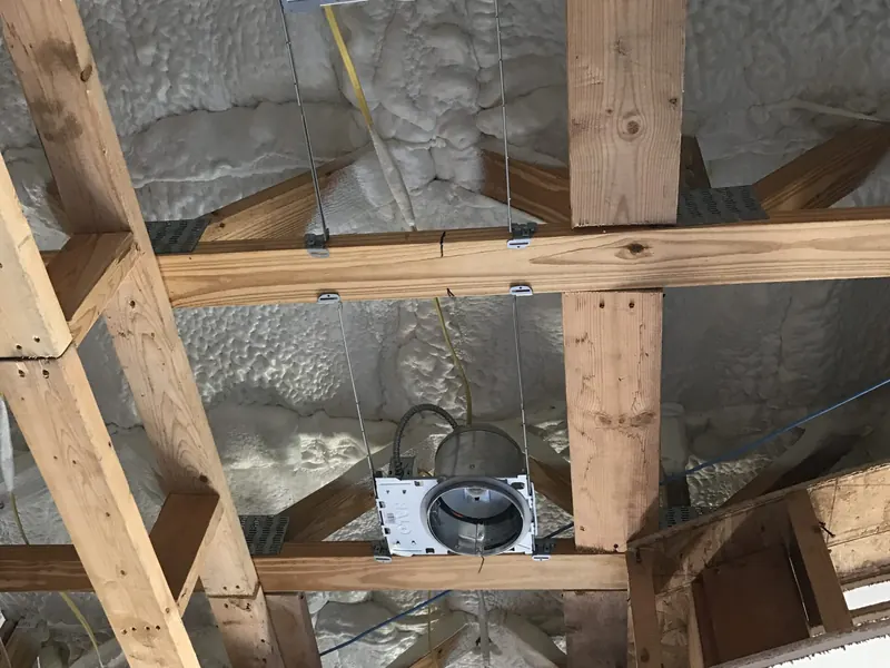 Attic framing insulated with spray foam around the roof deck, with flexible ductwork and a ceiling register running through the space