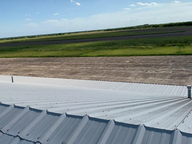 A coated metal roof edge in the foreground with green farm fields stretching out behind under a blue sky