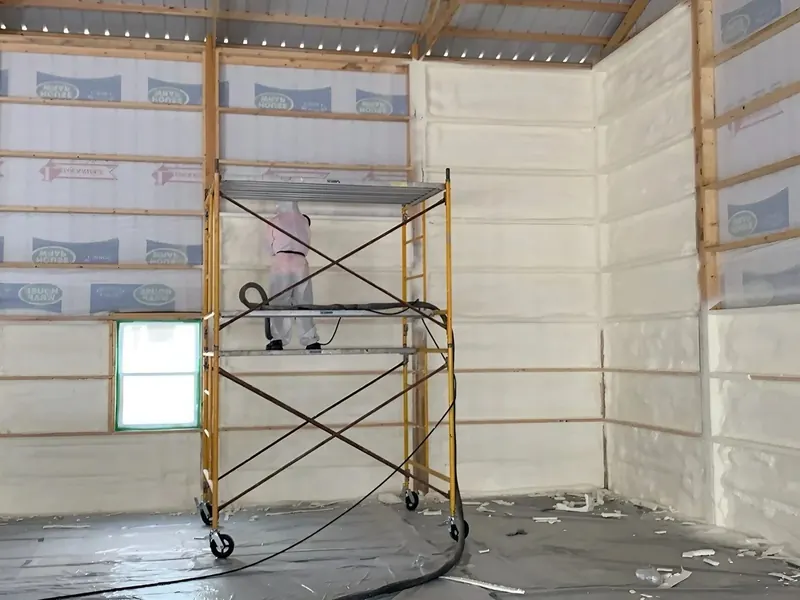 A commercial building interior with walls fully covered in white spray foam, a yellow rolling scaffold tower parked mid-room