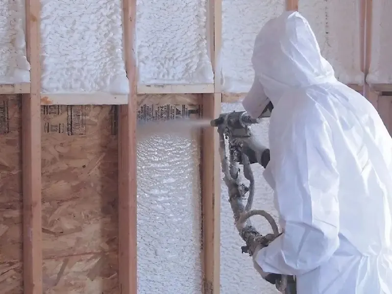 A Panda Insulation applicator in full white protective coveralls and respirator sprays open-cell foam into a stud cavity, the wood framing already filled with white foam