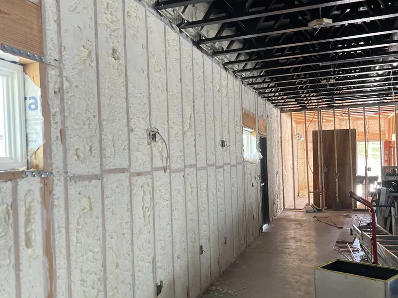 A long interior corridor with both walls insulated in white spray foam, framing still exposed, opening to a doorway at the end
