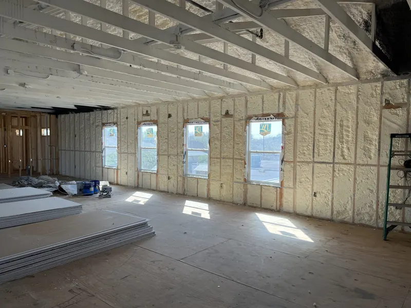 A wide interior insulated in white spray foam, sunlight falling through a row of windows onto stacked building materials