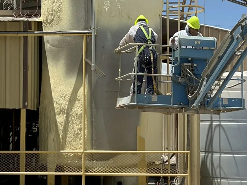 Two workers in hard hats and hi-visibility vests on the platform of a large industrial storage silo coated in protective insulation
