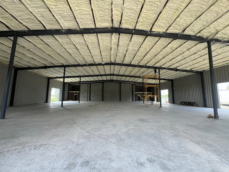 The interior of a steel-framed metal building after spray foam insulation, the entire underside of the roof deck covered in tan foam between the purlins
