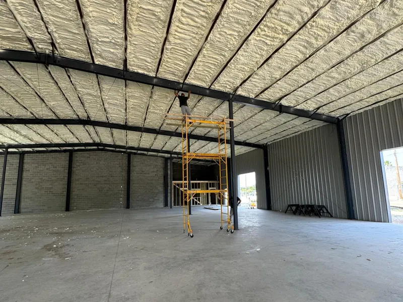 A steel building interior with the roof deck insulated in tan spray foam, the scaffold still standing at the far wall