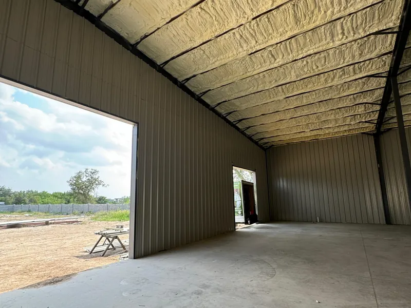 A metal building interior insulated overhead in spray foam, with the roll-up door open to daylight and open ground beyond