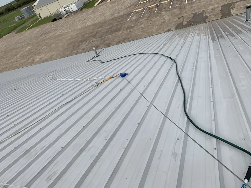 A spray wand and hose laid across a metal roof mid-coating, the near half already covered in fresh white coating