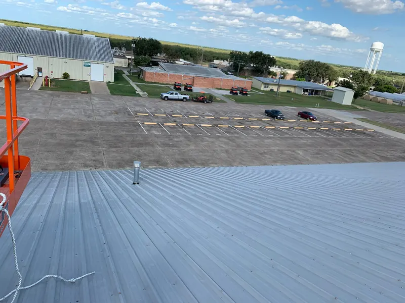 A coated standing-seam metal roof photographed from a raised boom lift, with the building parking lot, service vehicles and surrounding town visible below